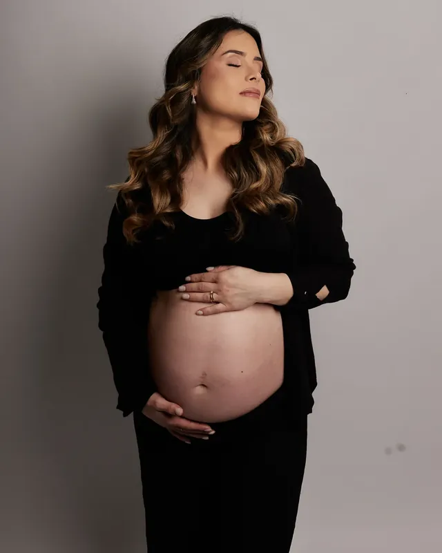 Gestante de olhos fechados com as mãos na barriga em ensaio de gestante em estúdio, em Curitiba