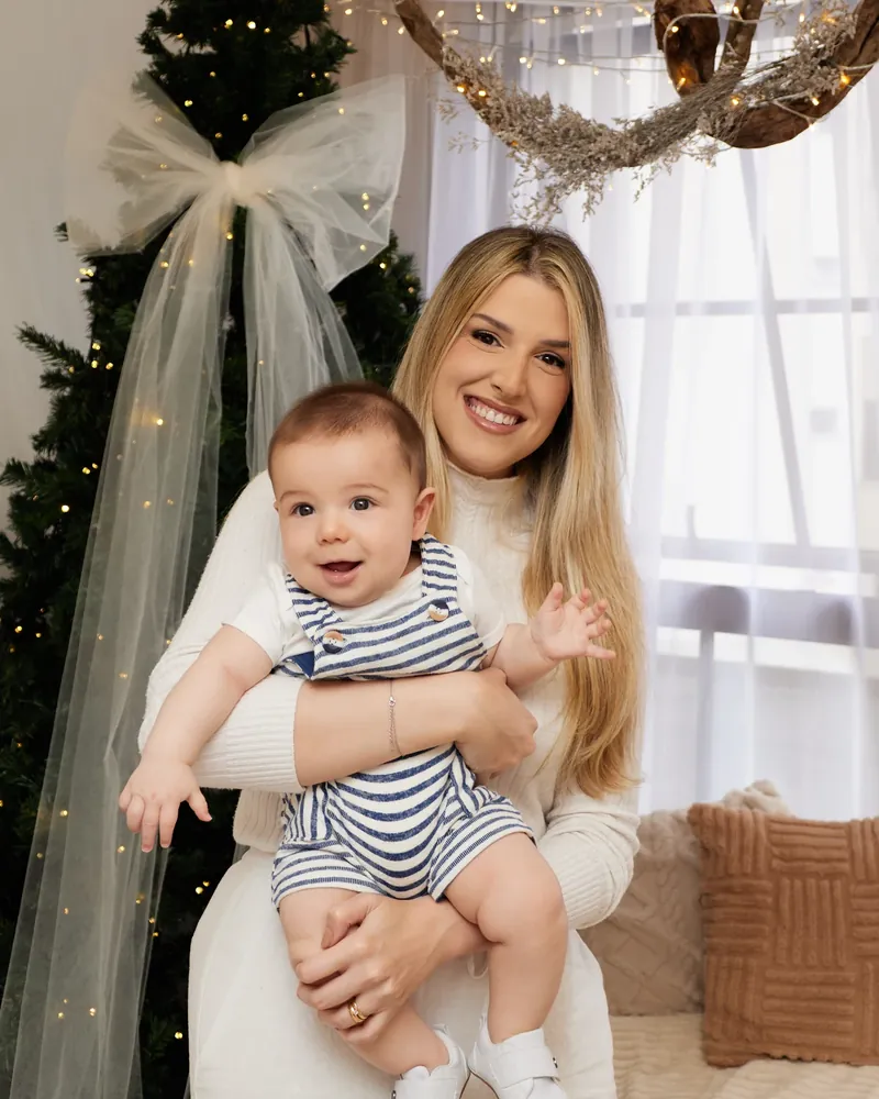 Mãe sorrindo com o bebê no colo, diante da árvore de Natal com luzes e cortina de tule, no cenário de Natal do estúdio