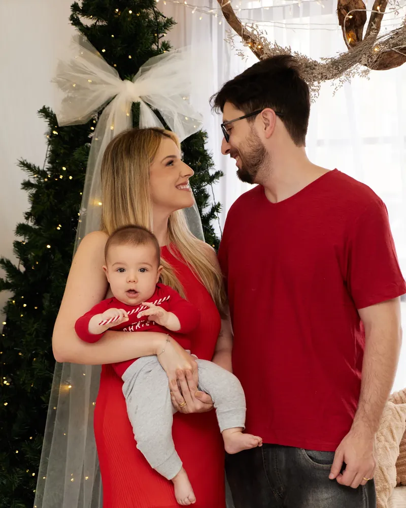 Família de roupas vermelhas com o bebê no colo, diante da árvore de Natal, no cenário do estúdio