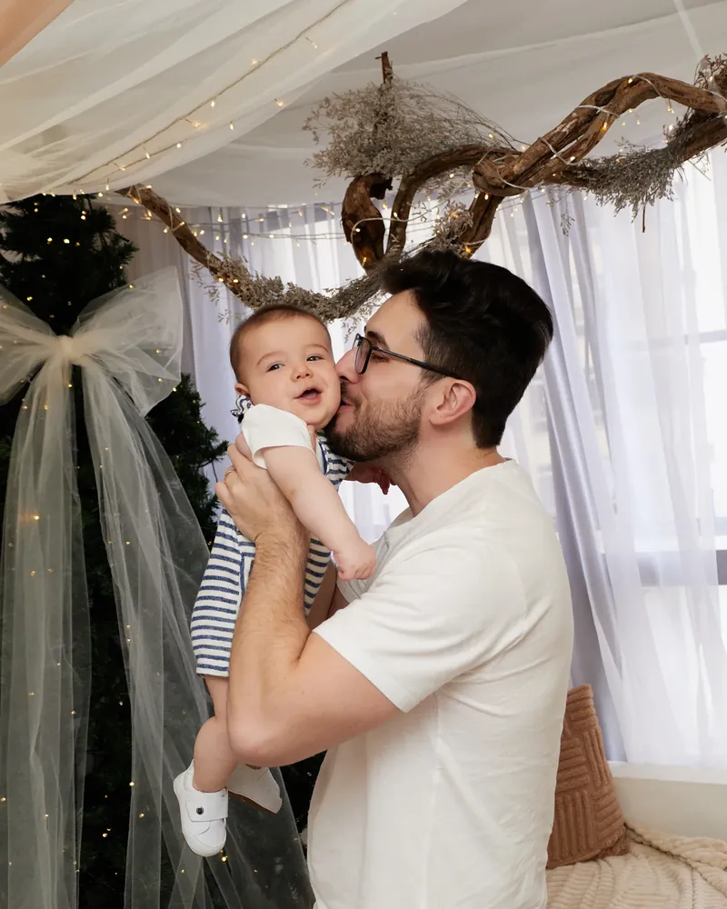 Pai beijando o bebê, diante da árvore de Natal e do tule iluminado, no cenário de Natal do estúdio