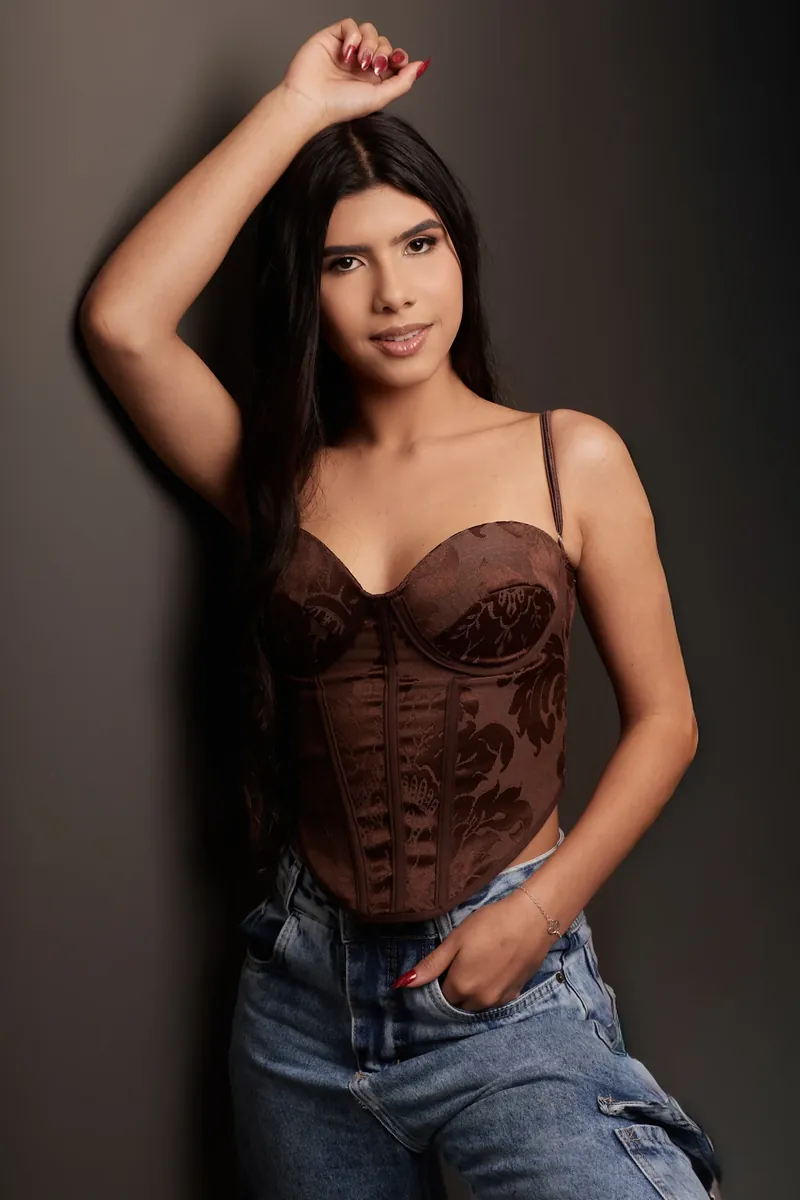 Mulher de cabelo escuro com o braço sobre a cabeça, de corpete marrom e calça jeans, em retrato no estúdio com fundo escuro