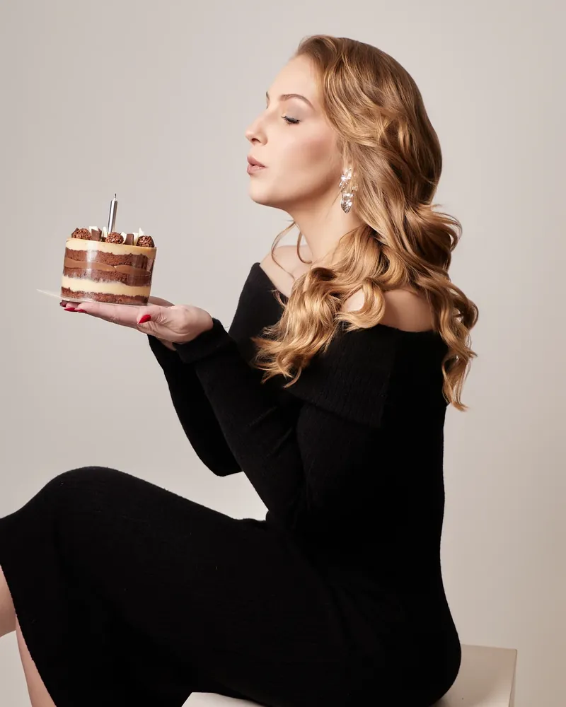 Mulher de vestido preto soprando a vela de um mini bolo, sentada em um cubo branco, em ensaio de aniversário