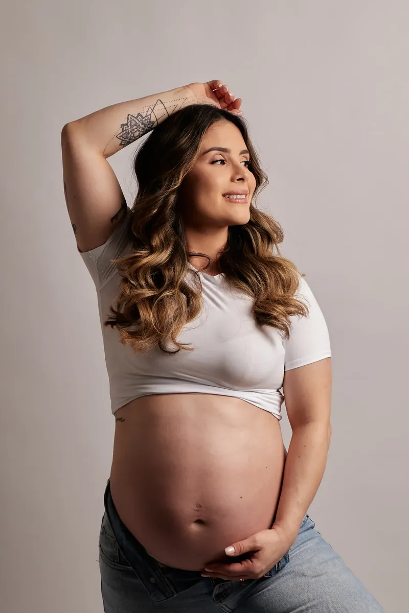 Gestante de camiseta branca e calça jeans com a mão sob a barriga, em ensaio de gestante no estúdio