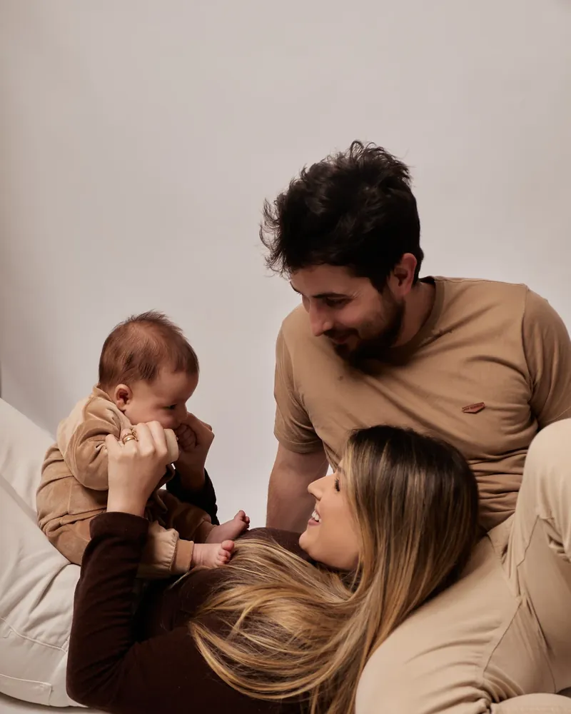 Mãe deitada com o bebê no colo e o pai ao lado, brincando, em ensaio de família no estúdio