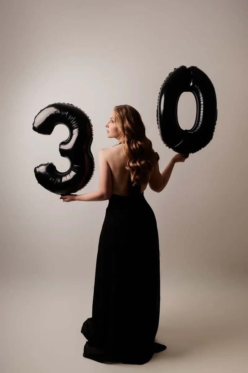 Mulher de costas, de vestido longo preto, entre dois balões pretos em forma de 3 e 0, em ensaio de aniversário de 30 anos